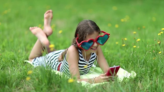Girl in glasses lies on grass and communicates via smartphone. Pretty girl in sunglasses with red smartphone lies on green grass. Female with phone. Girl with smartphone communicates through skype