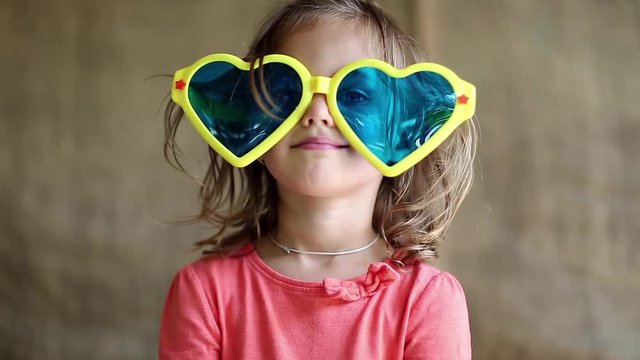 Funny little girl in big glasses looks at camera, shows his hands and tongue. Little girl in big sunglasses in the shape of hearts looks at the camera