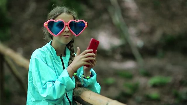 Girl in big sunglasses in the shape of hearts uses his smartphone. Girl with glasses makes selfie on his phone. Pretty girl with red cell phone makes photos. Female uses red smartphone