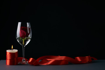Wineglass with rosebud inside, candle and red ribbon on dark background. Love card concept with copy space. Valentine's day theme