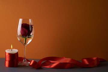 Wineglass with rosebud inside, candle and red ribbon on orange background. Love card concept with copy space. Valentine's day theme