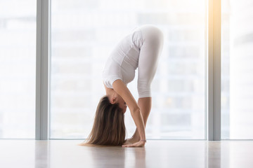 Young woman practicing yoga, standing in head to knees, uttanasana exercise, Standing forward bend pose, working out, wearing sportswear, white t-shirt, pants, full length, floor window