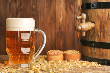 Glass of beer with ingredients and barrel on wooden background