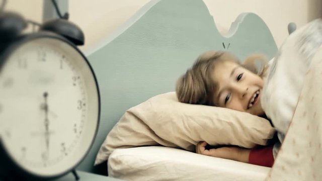 A little girl sleeping in her bed. The alarm clock near her is about to ring. She gladly wakes up. Blurred on the left side.
