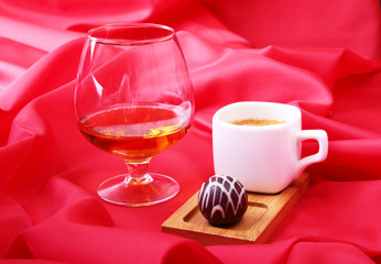 White cup of coffee and cognac in a glasses, pralines on red background.
