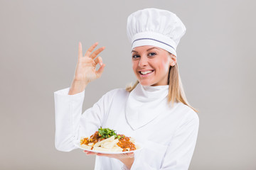 Beautiful female chef is showing ok sign and prepared meal on gray background.
