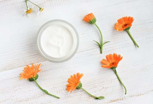 Cosmetic container of marigold soothing body care cream, fresh orange calendula flowers, white wooden table overhead view.