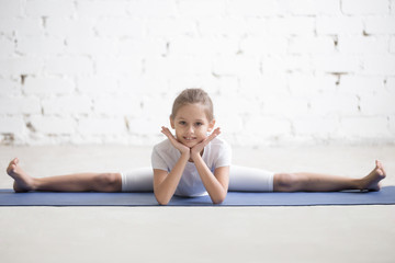 Funny portrait of cute girl child practicing yoga, doing splits, Samakonasana stretching exercise, Straight Angle pose, working out wearing sportswear, indoor full length, white loft studio background