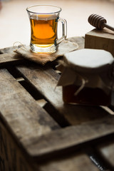 Glass cup with hot tea with jar of honey or jam, wood spoon, on  vintage box, rustic vintage interior,cozy atmosphere, back lighting