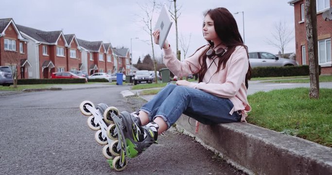 Pretty young girl in roller skates and headphones sitting on country street. Girl using tablet computer for selfie video. Ireland 2016. Red Epic 4k.