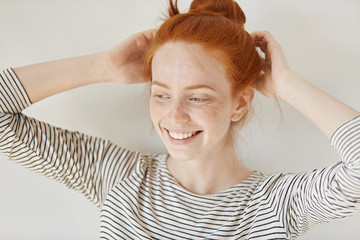 Fototapeta premium People and lifestyle concept. Attractive young woman with ginger hair and freckled skin wearing striped top smiling happily while adjusting her hairstyle before going out to party with her friends