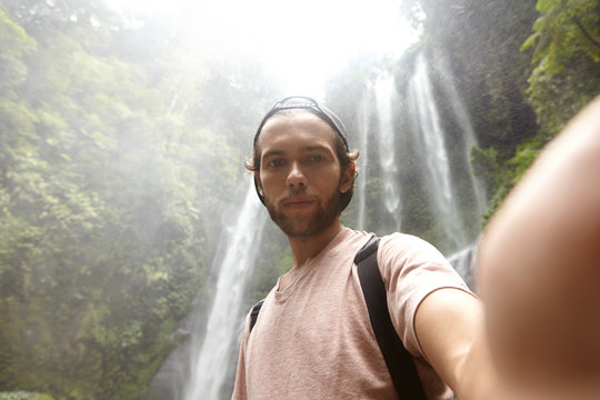 High-angle shot of handsome bearded male tourist taking selfie on front-facing camera of some electronic device, posing outdoors against amazing view background, river running down mountain behind him