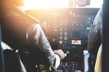 Two unrecognizable jet airliner pilots wearing leather jackets piloting aircraft at sunset, sitting inside cabin at steering control with modern dashboard, sun shining through cockpit windscreen - obrazy, fototapety, plakaty