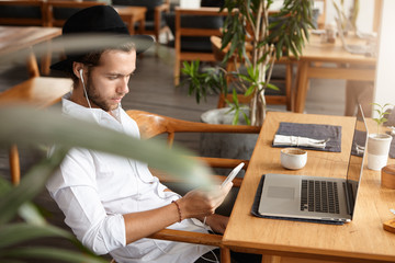 Side view of handsome young Caucasian freelancer or student sitting at cafe table with open laptop pc, holding mobile phone and listening to music on earphones, using online app during breakfast