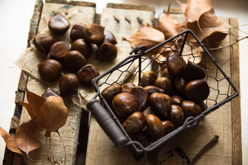 Fresh shiny chestnuts in wire basket scattered on burlap cloth, dry brown leaves,on vintage wood box, autumn, fall, harvest, cozy atmosphere, top view