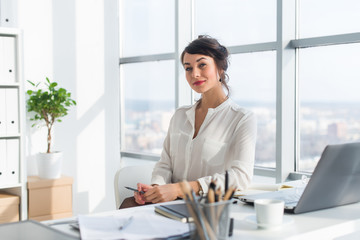 Close-up portrait of a woman sitting in modern loft office, smiling, looking at camera. Young confident female business worker ready for the work day.