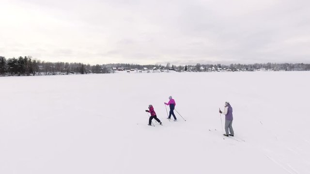 Back aerial view at three women running on ski on the ice of lake to the village on the shore. Camera flying
