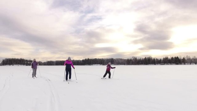 Grandmother, mother and young daughter skiing in the snowy ice of frozen lake at evening time. Russia. Camera going up