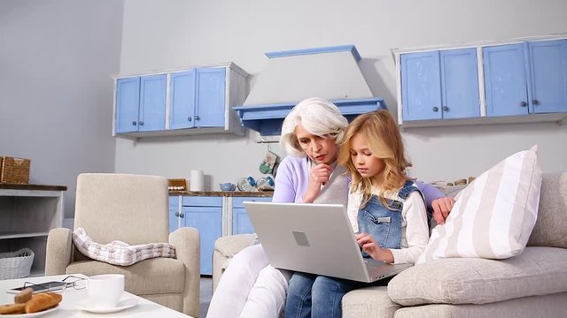 Granny and little girl using laptop computer at home all together. Little blond lady typing and her granny looking at screen. Happy girls expressing positive emotions.