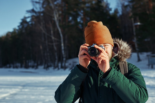 Close up portrait of photographer with vintage camera. Bearded backpacker making photo in sunny day contryside with snowy field and green forest. Travel concept with free space for advertising.