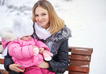 Mother holds daughter on hands outdoors frost. She sat on the bench. Family walk in a winter park. Family happiness.