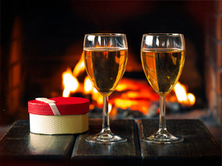Two glasses of wine and a gift box in the shape of a heart on the background of the fireplace
