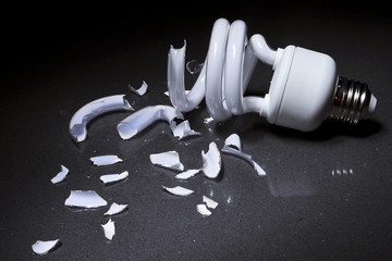 A Broken Compact Fluorescent CFL bulb lays shattered in shards of glass on a hard surface with a dark background