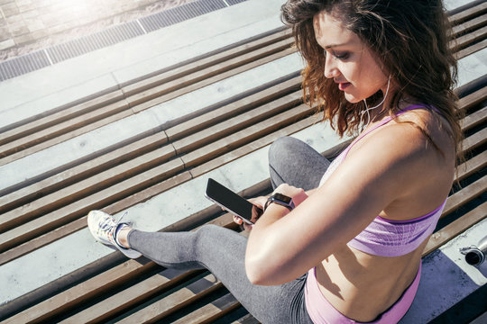 Summer sunny day. View from above. Young woman in sportswear sits outside and synchronizes fitness tracker with your smartphone. Girl is resting after training, looking at screen of smartwatch.