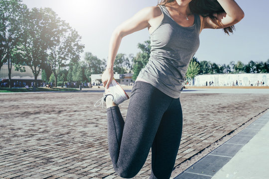 Cropped image. Summer sunny day. Young woman in gray sportswear standing outdoors and stretch before exercise. Girl stretches legs before jogging.
