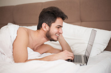 Young smiling man using laptop in bed at home