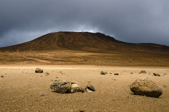 Desolate moonscape in the national park of Mount Kilimanjaro. Rocks lay a moon like landscape extending to a hill that is surrounded by dark clouds. 