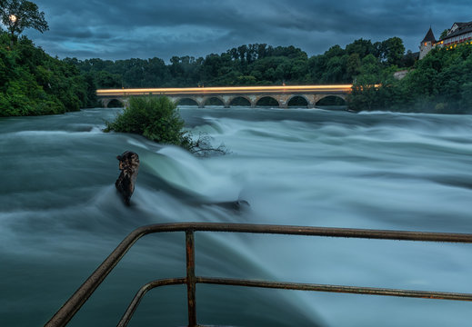 Long night exposure of the Rhine River right before the Rhine Falls. A train leaves its light trails behind on the bridge in the back. A railing is in the foreground on the waters edge.