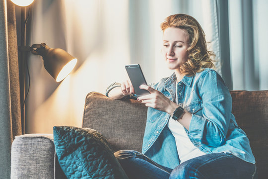 Young smiling woman in denim shirt sitting at home on couch and using smartphone. Girl uses digital gadget. Female online shopping, surfing internet, chatting, blogging. Freelancer working home.