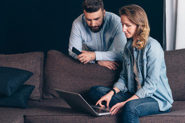 Young woman in denim shirt,sitting at home on couch and using laptop.Guy holding smartphone.Couple shopping online,surfing internet.