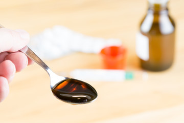 Ein Teelöffel mit dunklem Hustensaft, einnehmen, im Hintergrund die Flasche mit Medizin und ein Fieberthermometer