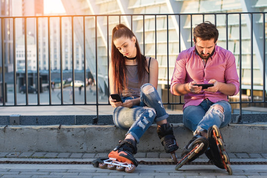 Couple with phones sitting. Inline skaters on urban background. Pay attention to your partner.