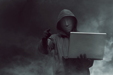 Portrait of hooded hacker with mask holding laptop while standing