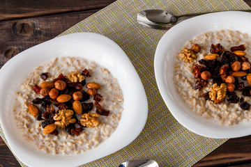 Two plates of homemade oatmeal porridge with dried plums, almonds, hazelnuts, walnuts and dates on old dark grey rustic wooden table with spoon. Hot and healthy food for Breakfast.
