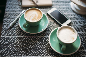 Fresh cup of cappuccino in a green cup on a coffee table with a smartphone and a notebook