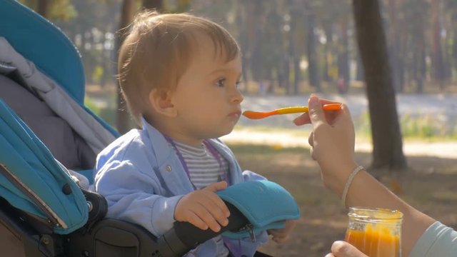 Closeup portrait of cute baby boy sitting in stroller at park and eating apple sauce