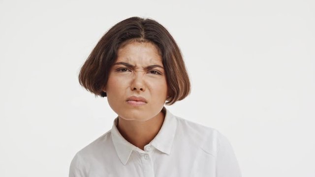 Young beautiful brunette east asian female in shirt looking suspiciously at camera showing disapproval with two thumbs on white background in slowmotion