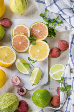 Variety of whole and sliced citrus fruits pink tiger lemon, lemon, lime, mint and exotic lichee on white chopping board with kitchen towel over white concrete textured background. Top view. Healthy