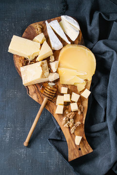 Cheese plate. Assortment of cheese with walnuts, honey from honey dipper on olive wood serving board with textile over dark blue canvas as background. Top view with space.