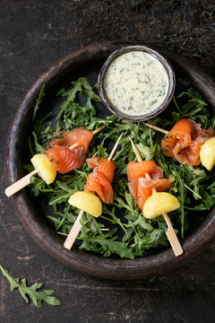 Delicious Appetizer with smoked salted salmon and boiled potatoes on skewers served with creamy dill sauce and arugula in stone plate over dark background. Top view, copy space