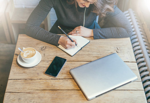 Young business woman sitting at wooden table in cafe and use smartphone while making notes in notebook. Nearby is cup of coffee and closed notebook.