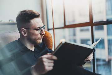 Attractive stylish businessman reading black book while sitting in vintage chair at his modern office.Young man relaxing after work day.Panoramic windows on blurred background.Horizontal,film effect.