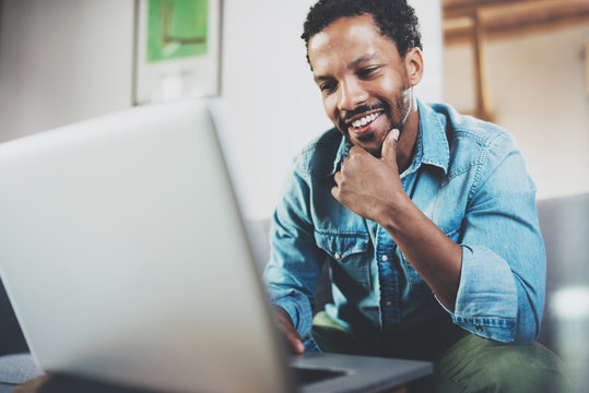 Smiling young African man making video call with friends while sitting on sofa at his modern office.Concept of happy business people.Blurred background, flare effect.