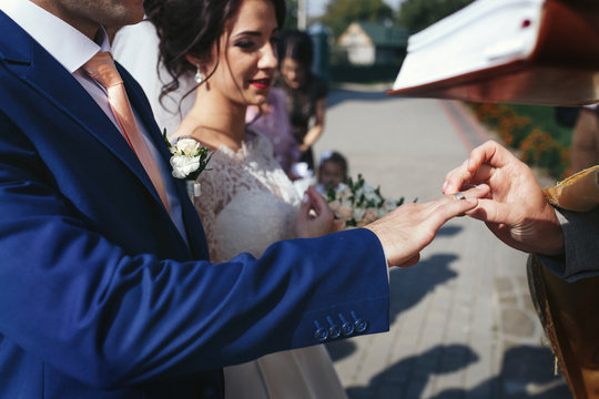 Priest puts wedding ring on groom's finger while they stand outs