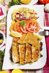 Breaded and deep fried hake fillets on a white plate with lemon, tomatoes and potato salad.