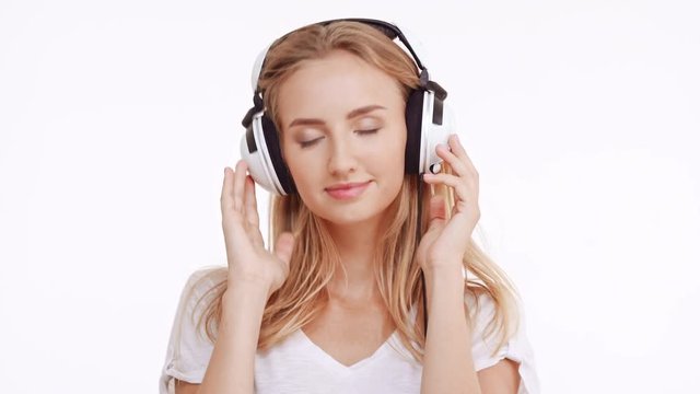 Young beautiful Caucasian blonde girl listening music through headphones dancing smiling singing on white background
